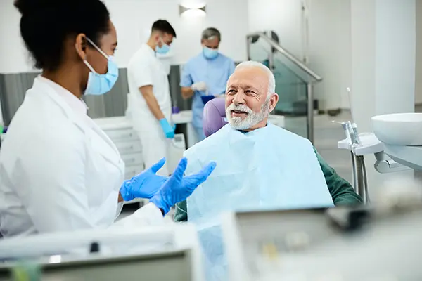 Older white patient sitting in dental chair calmly discussing his oral cancer screening with his Black female dentist