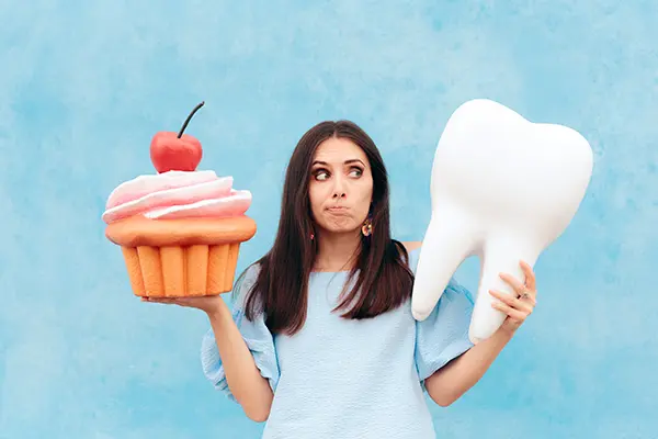 An undecided woman holds a giant tooth model in one hand and a giant cupcake in the other, metaphorically weighing the effect of a poor diet will have on her dental health.